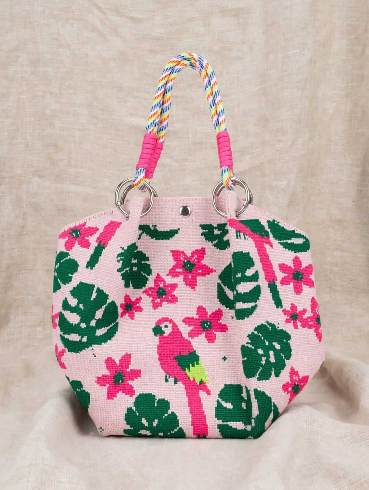 A pink woven tote bag with a tropical print featuring green monstera leaves, pink flowers, and a pink parrot, complete with colorful braided rope handles and silver metal rings, set against a neutral linen background.