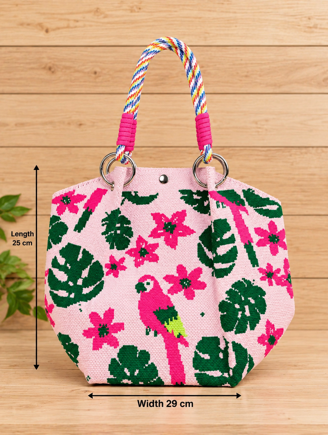 A pink woven tote bag with a tropical print featuring green monstera leaves, pink flowers, and a pink parrot, complete with colorful braided rope handles and silver metal rings, set against a neutral linen background.