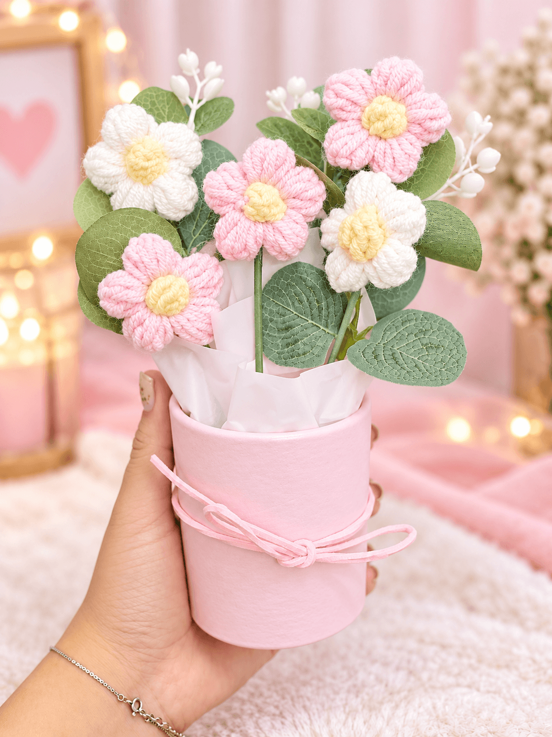 Five pink and white crochet-style flowers with yellow centres, decorative green leaves, white floral accents and wrapping arranged in a pastel pink cylindrical gift box.