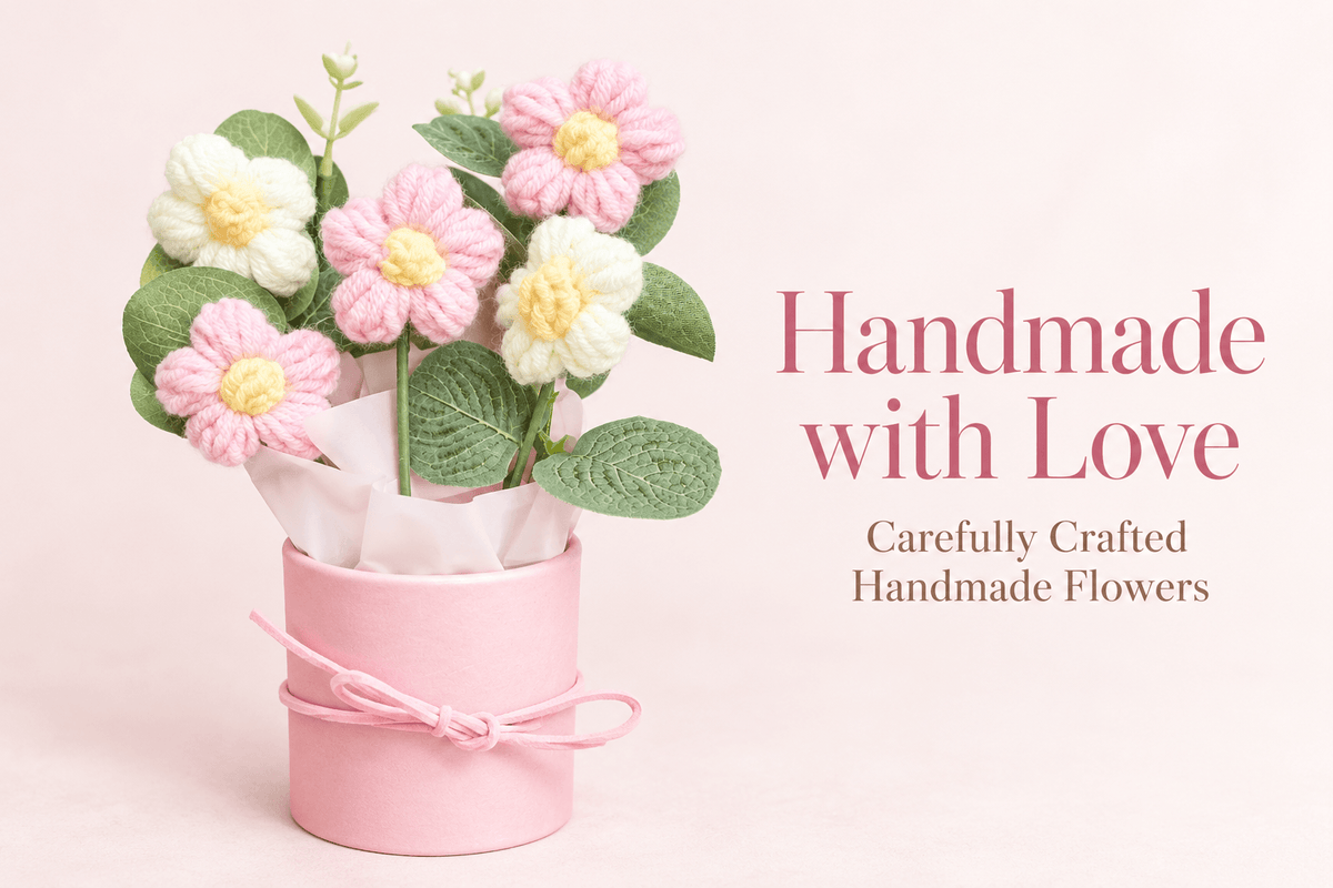 Five pink and white crochet-style flowers with yellow centres, decorative green leaves, white floral accents and wrapping arranged in a pastel pink cylindrical gift box.