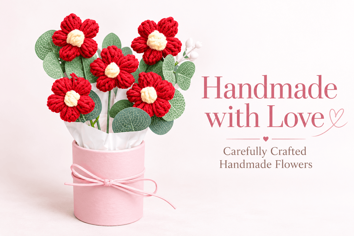 Five red crochet-style flowers with cream-white centres, decorative green leaves and white wrapping arranged in a pastel pink cylindrical gift box with ribbon.