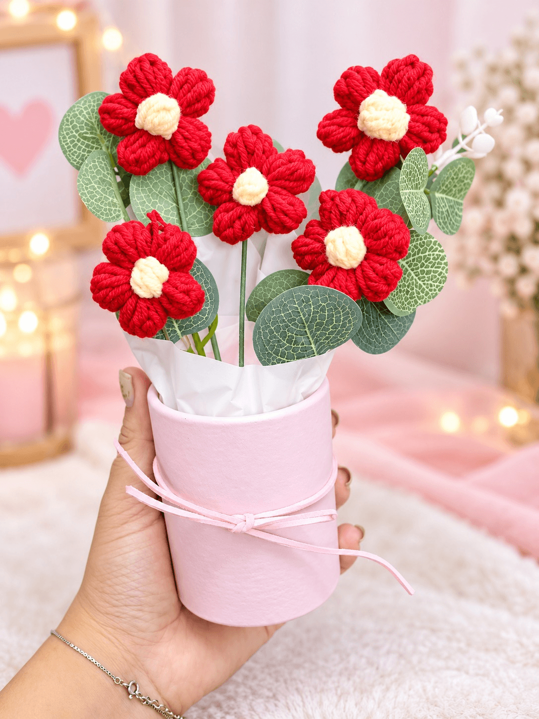 Five red crochet-style flowers with cream-white centres, decorative green leaves and white wrapping arranged in a pastel pink cylindrical gift box with ribbon.