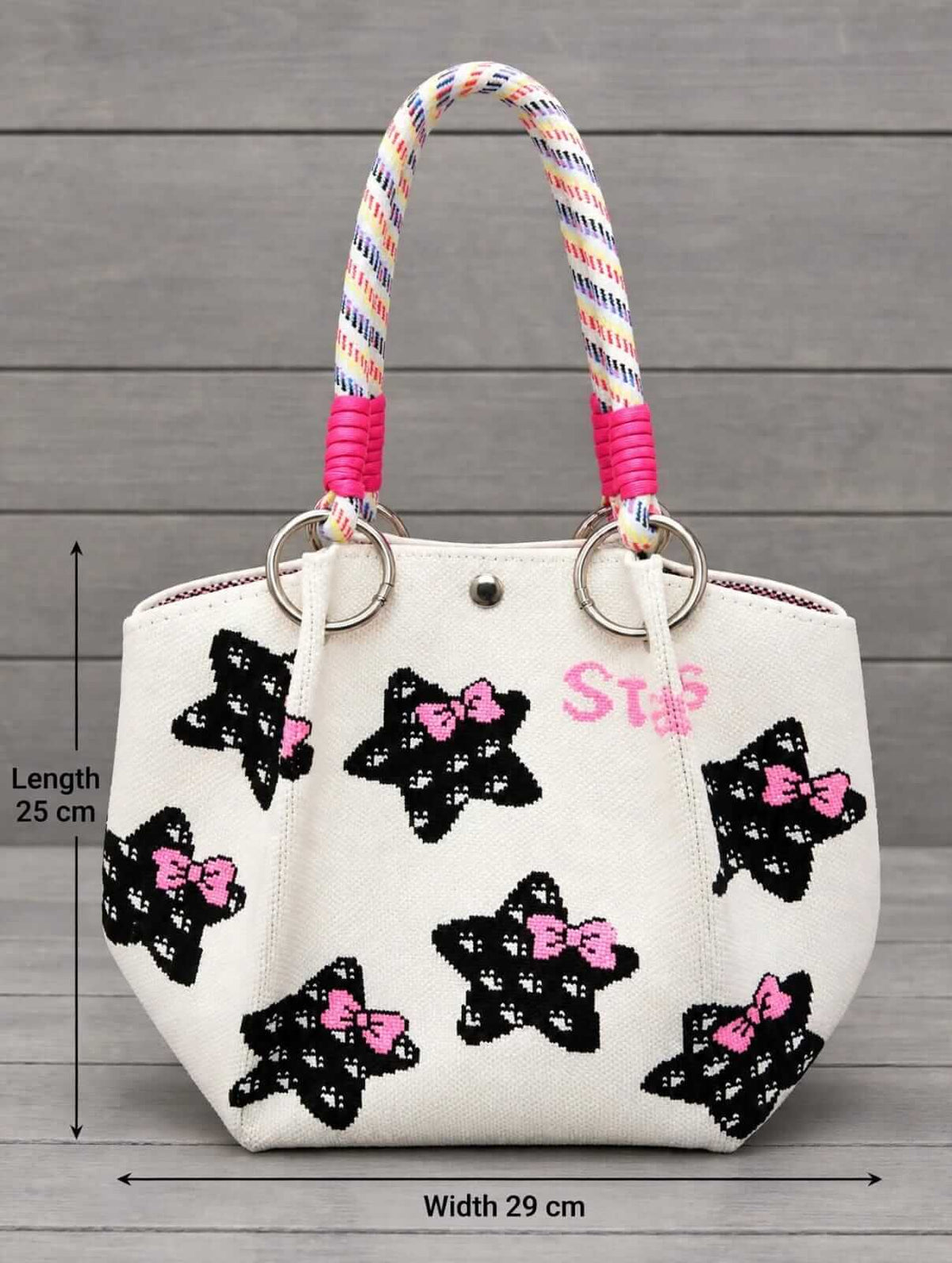An off-white canvas tote bag with black sequined star motifs decorated with pink bows, featuring multicolored striped rope handles and pink rope accents. The image includes dimension lines indicating a length of 25 cm and a width of 29 cm, set against a gray wood plank background.