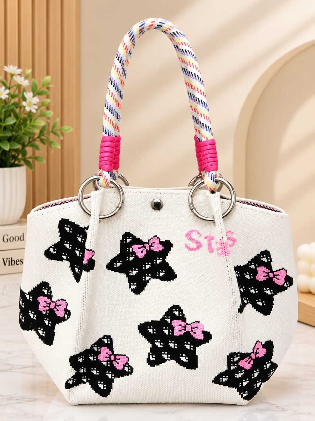 A white canvas tote bag decorated with black star motifs that have pink bows, featuring a colorful braided rope handle and pink lettering on the front.